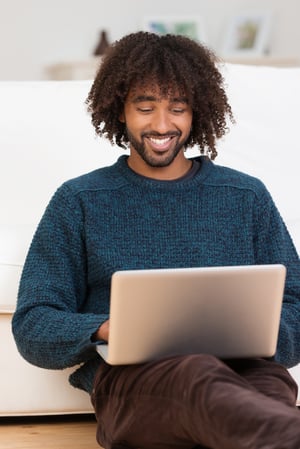 Handsome hip young African American man smiling as he types on his laptop computer while relaxing on the floor in the living room