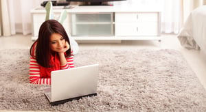 Portrait of dreamy smiling young woman using laptop while lying on floor at home