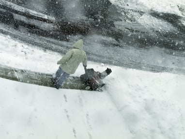 Winter at a glance Unrecognizable man in hooded coat stands holding a snow blower while he looks downward and snow falls all around him | Tips for Finding a Temporary Holiday Job